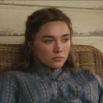 "A young woman in a blue patterned dress sits pensively in a wicker chair against a worn wooden wall, appearing thoughtful and somber in a rustic, vintage setting."