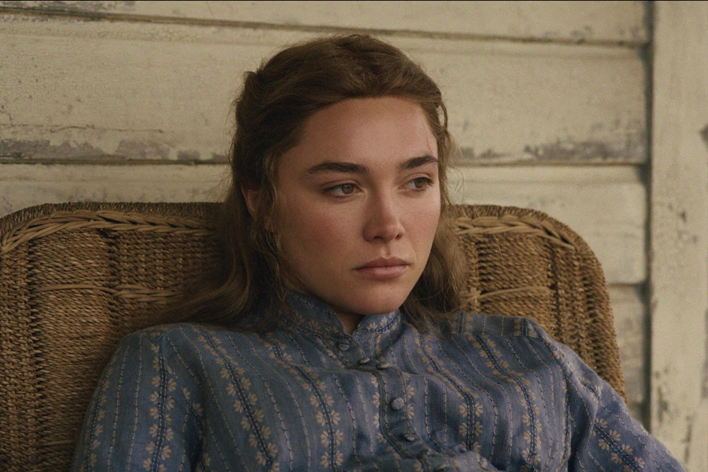 "A young woman in a blue patterned dress sits pensively in a wicker chair against a worn wooden wall, appearing thoughtful and somber in a rustic, vintage setting."